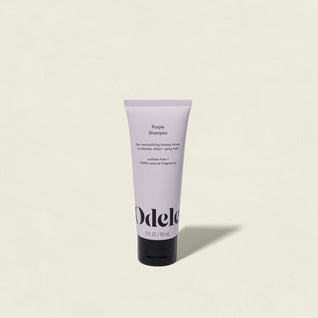 A tube of Purple Shampoo Mini stands upright against a plain, off-white background. This lavender-colored shampoo is designed for blonde hair, helping to neutralize brassy tones and create a brighter appearance.