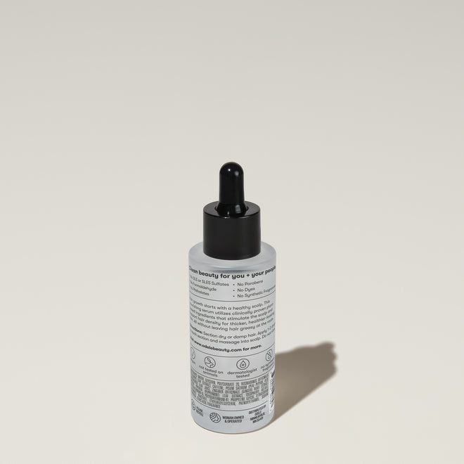 A bottle of Odele Rejuvenating Scalp Serum stands upright against a plain, light-colored backdrop with the back of the label facing the camera.