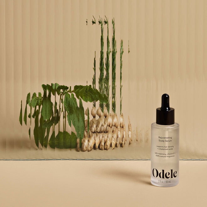 A bottle of Odele Rejuvenating Scalp Serum stands on a beige surface in front of a wall of mirrors that reflect mint, ginger and rosemary.