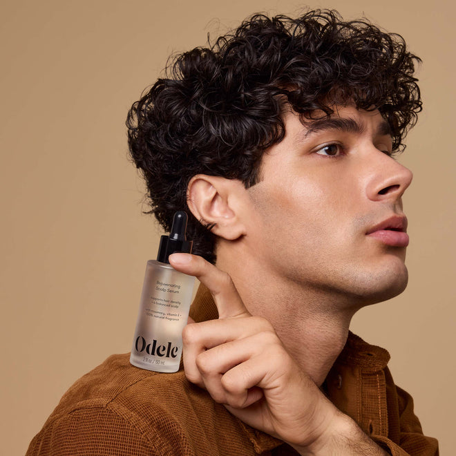 A young man with curly dark hair holds a bottle of Odele Rejuvenating Scalp Serum on their shoulder in front of a light orange background.