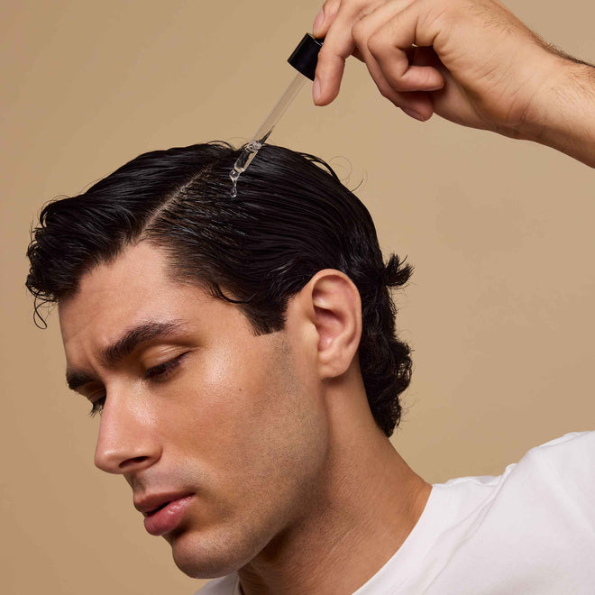 A man in a white shirt with dark, slicked-back hair applies Odele Rejuvenating Scalp Serum with a dropper, focusing on his scalp.