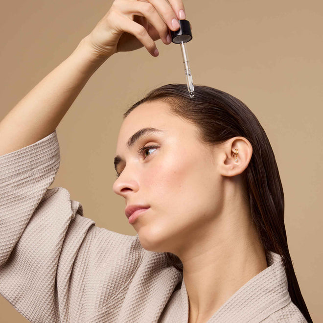 A young woman in a beige robe applies Odele Rejuvenating Scalp Serum to her scalp in front of a beige background.