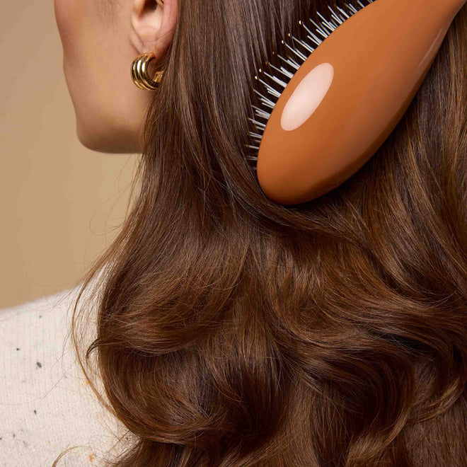 A woman with wavy brown hair, gold hoop earrings, and a light sweater brushes her hair with an orange brush.
