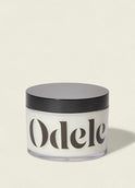 A clear jar with a black lid, filled with white cream and labeled Odele, sits against a plain light background—highlighting the Scalp + Body Scrub's salon-quality moisturizing formula.