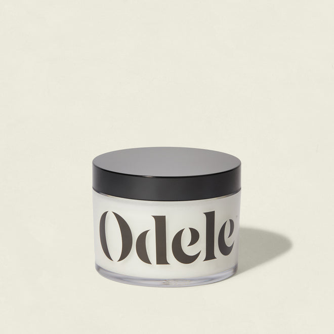 A clear jar with a black lid, filled with white cream and labeled Odele, sits against a plain light background—highlighting the Scalp + Body Scrub's salon-quality moisturizing formula.