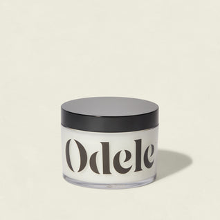 A clear jar with a black lid, filled with white cream and labeled Odele, sits against a plain light background—highlighting the Scalp + Body Scrub's salon-quality moisturizing formula.