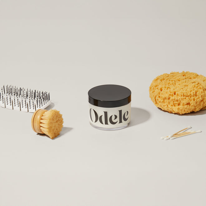 A jar of Odele Scalp & Body Scrub sits on a light surface, surrounded by a round body brush for exfoliation, a bath sponge, hairbrush, and cotton swabs.