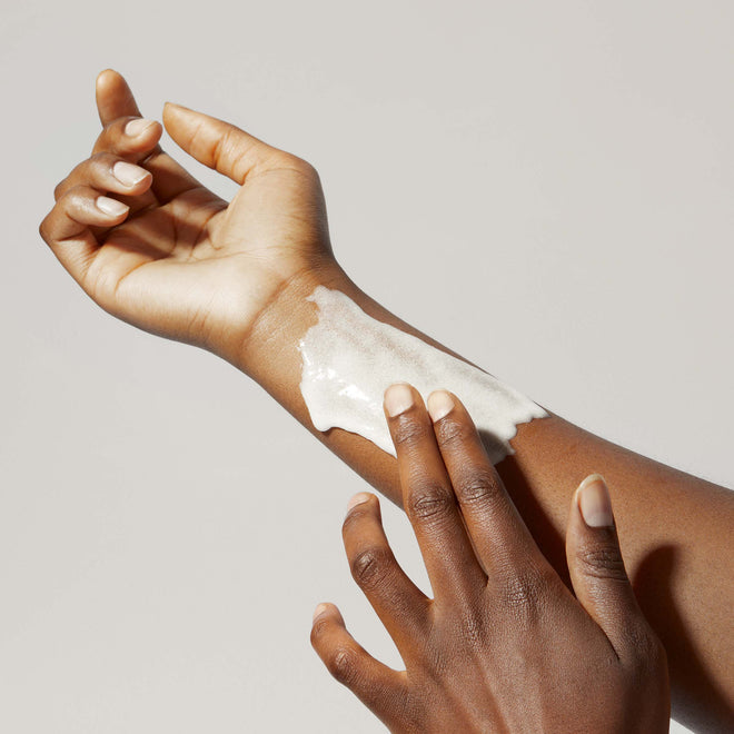 A person with medium brown skin applies Odele Scalp & Body Scrub to their forearm with their fingers against a plain, light background.
