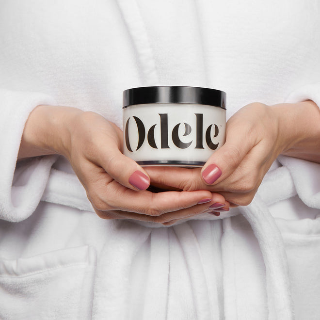 A person in a white robe holds a jar of Odele Scalp & Body Scrub in front of their midsection.