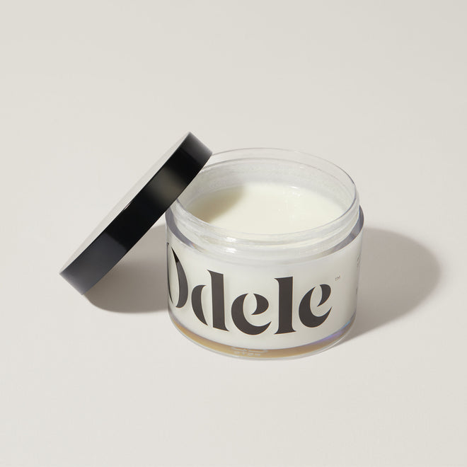 An open jar of Odele Scalp & Body Scrub sits on a plain light-colored surface with its lid leading against the ege of the jar.