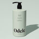 A light green bottle of Odele Smoothing Shampoo Bulk with a black pump, featuring benefits like extra moisture, frizz control, shine, sulfate-free formula, and natural fragrance, set against a pale green background.