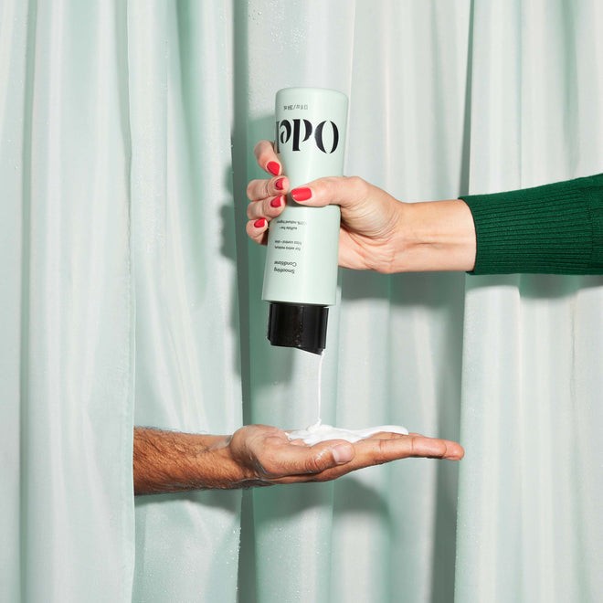 A person in a green sleeve dispenses Odele Smoothing Conditioner onto another’s open palm, both arms emerging from pastel green shower curtains.