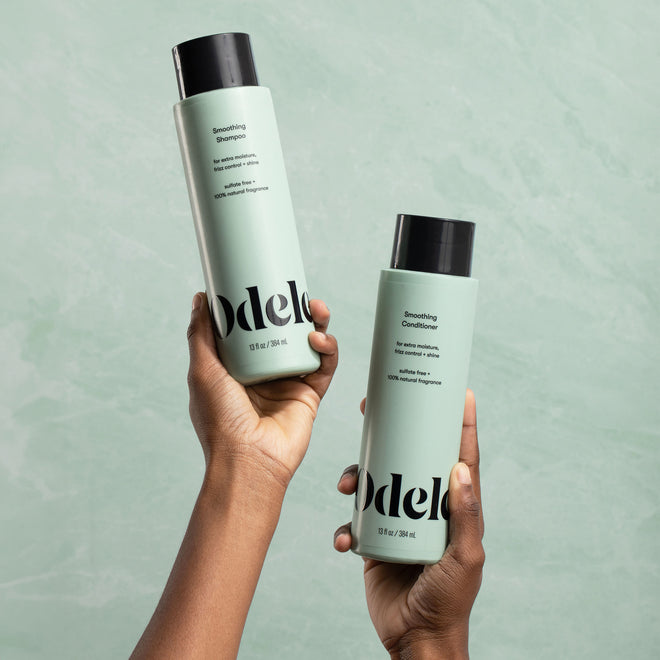 Two hands hold Odele Smoothing Shampoo and Conditioner in front of a light green background.