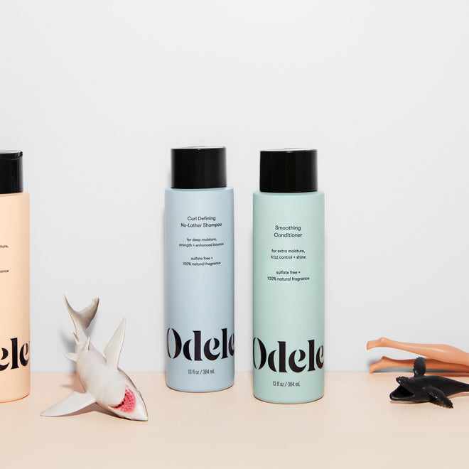 Three Odele bottles stand on a plain light surface surrounded by scattered children's toys.