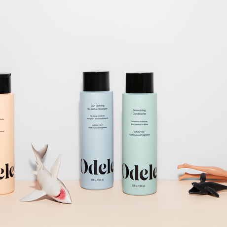 Three Odele bottles stand on a plain light surface surrounded by scattered children's toys.