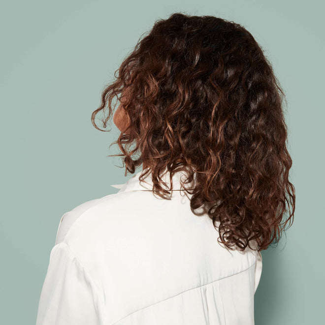 A young woman with shoulder-length brown waves wears a white shirt and is shown from behind against a plain light green background.