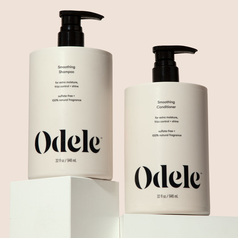 Two bottles of Odele Smoothing Shampoo and Conditioner are displayed on white blocks in front of a plain light-colored background.