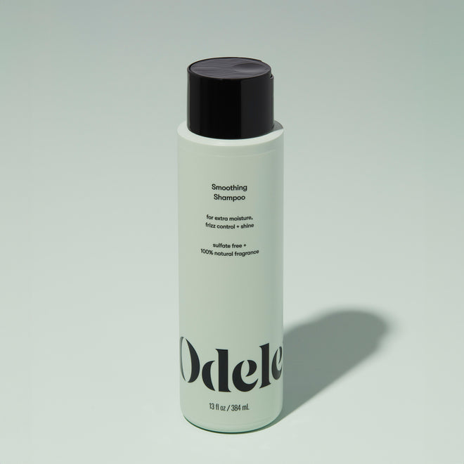 A bottle of Odele Smoothing Shampoo stands upright against a plain light green backdrop with the front of the label facing the camera.