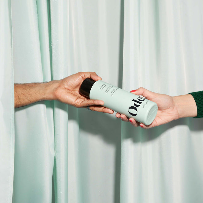Two hands, one from each side, exchange a bottle of Odele Smoothing Shampoo in front of matching curtains.