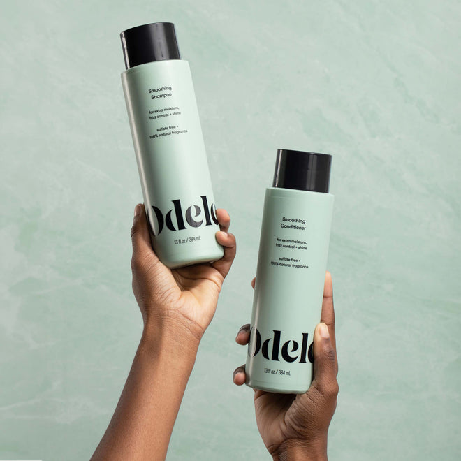 Two hands hold up bottles of Odele Smoothing Shampoo and Smoothing Conditioner in front of a plain light green background.