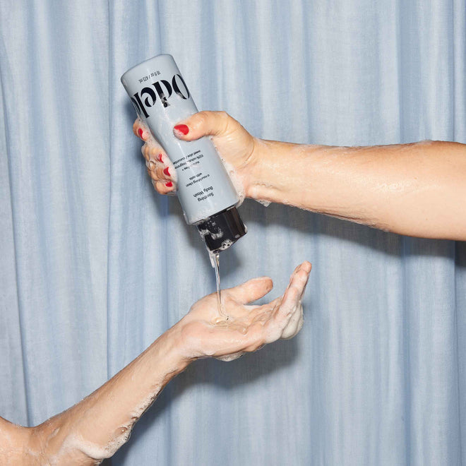 A hand squeezes Odele Soothing Body Wash into another person’s soapy hand, set against a light blue curtain background.