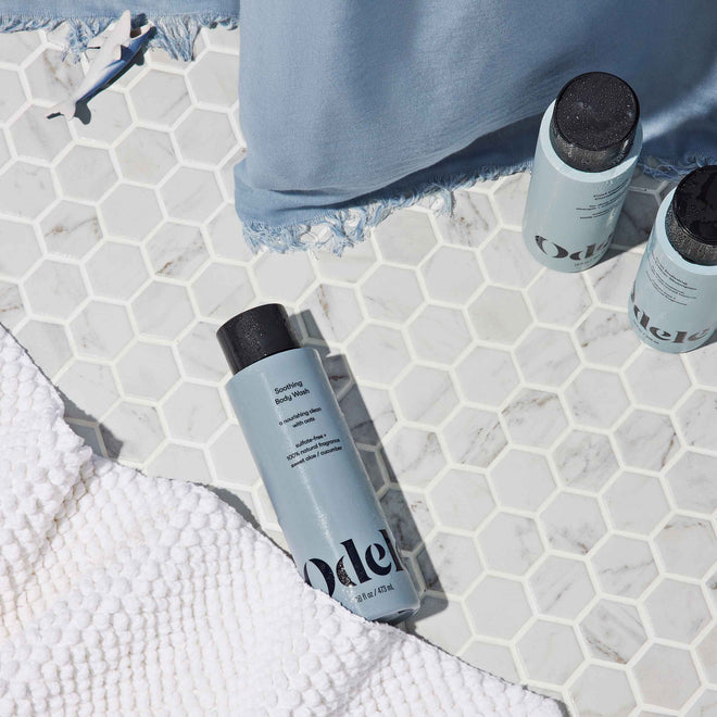 Light blue bottles of Odele Soothing Body Wash rest on white hexagon tile with a blue curtain and a textured white towel, creating a clean, spa-like vibe.