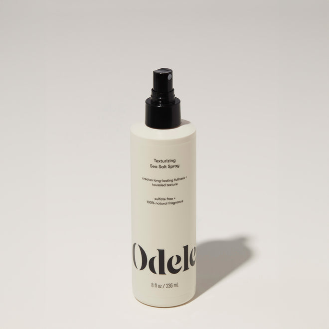 A bottle of Odele Texturizing Sea Salt Spray stands upright against a plain, light-colored backdrop with the front of the label facing the camera.