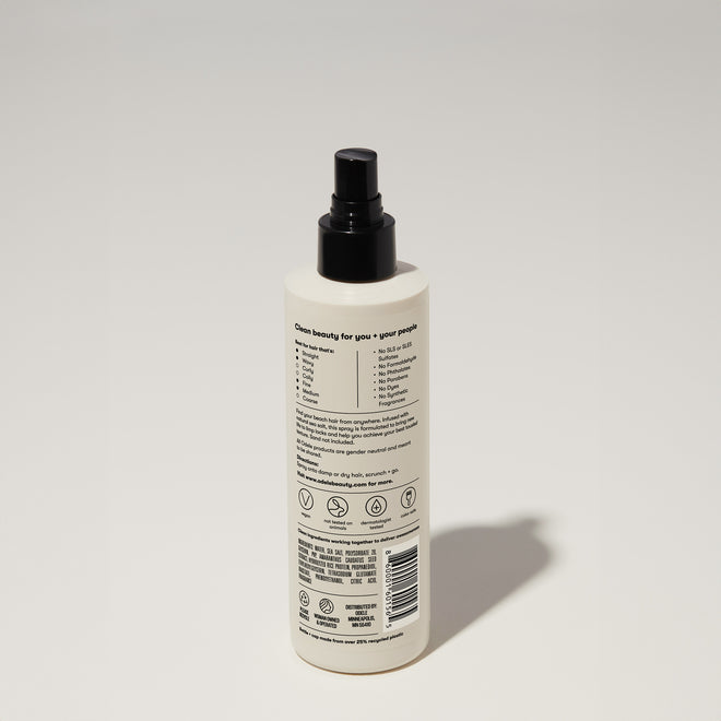 A bottle of Odele Texturizing Sea Salt Spray stands upright against a plain, light-colored backdrop with the back of the label facing the camera.