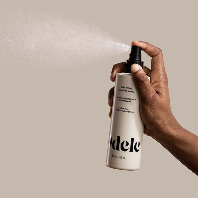 A brown-skinned hand sprays Odele Texturizing Sea Salt Spray in front of a plain light beige background.