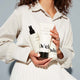 A woman in a white blouse and skirt holds three Odele products with both hands against a plain, light blue background. Her face is not shown.