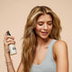 A young woman with long, wavy blonde hair smiles while spraying Odele Texturizing Sea Salt Spray onto her hair in front of a plain light orange background.
