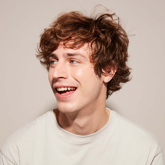 A smiling young man with short wavy red-brown hair wears a white t-shirt and looks off to the side in front of a plain light colored background.