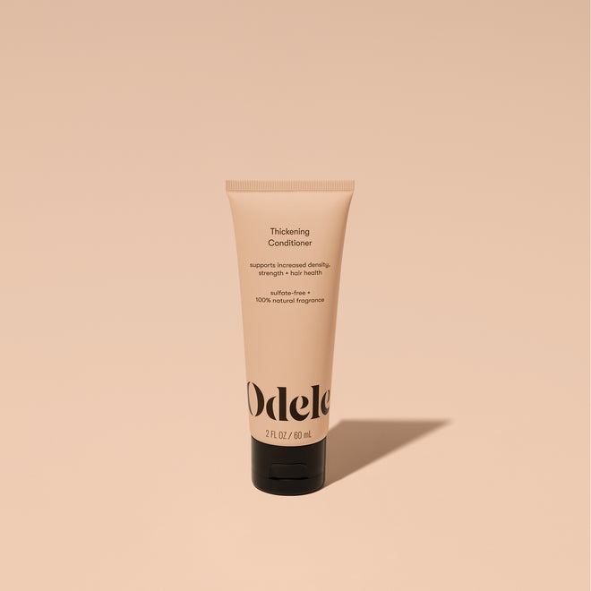 A tube of Odele Thickening Conditioner stands upright against a plain, light orange backdrop with the front of the label facing the camera.