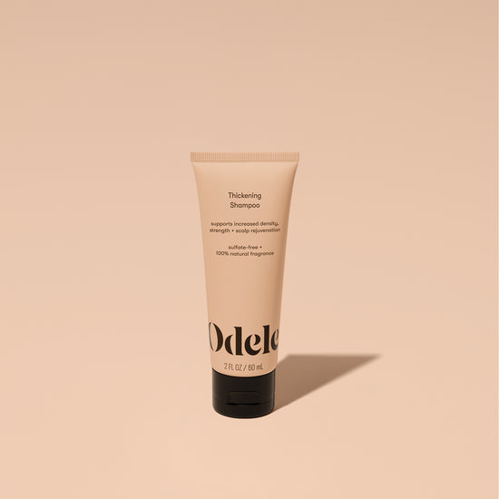 A tube of Odele Thickening Shampoo stands upright against a plain, light orange backdrop with the front of the label facing the camera.