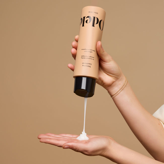 A hand dispenses Odele Thickening Conditioner onto an outstretched palm against a plain beige background.