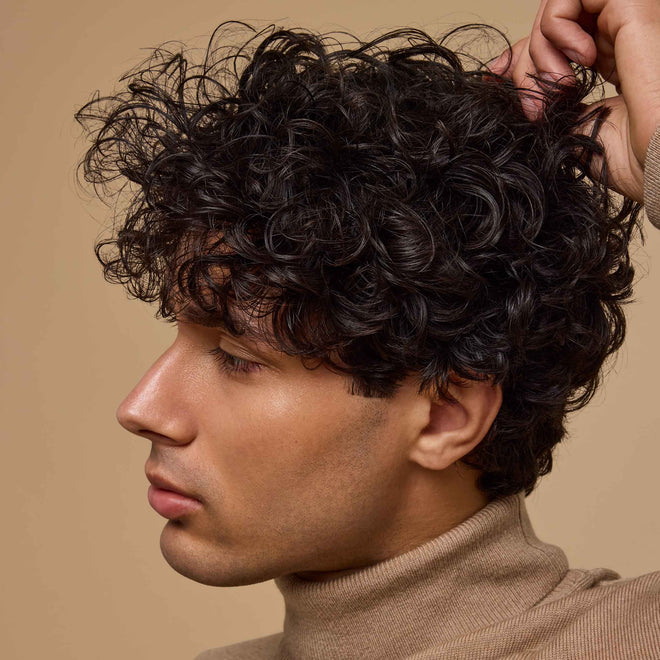 A young man with short curly dark hair wears a beige turtleneck and gently touches his hair while looking off to the side in front of a neutral tan background.