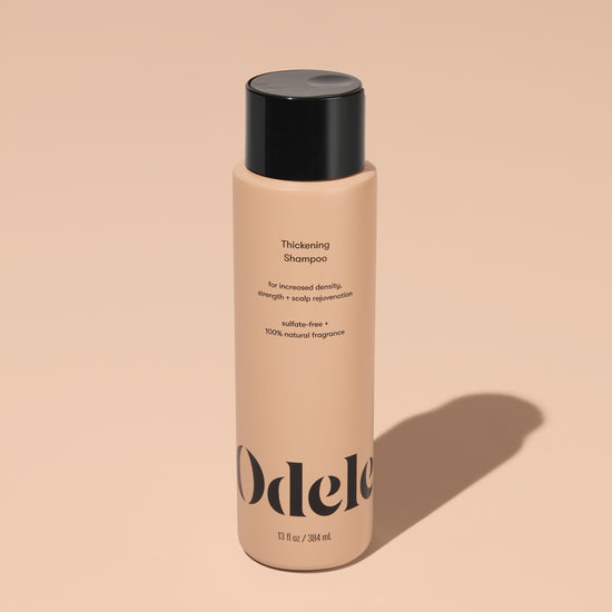 A bottle of Odele Thickening Shampoo stands upright against a plain, light orange backdrop with the front of the label facing the camera.