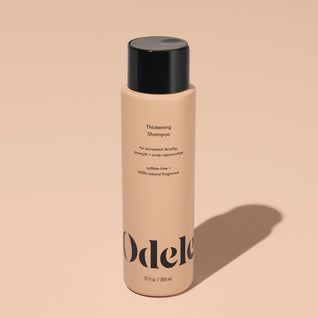 A bottle of Odele Thickening Shampoo stands upright against a plain, light orange backdrop with the front of the label facing the camera.