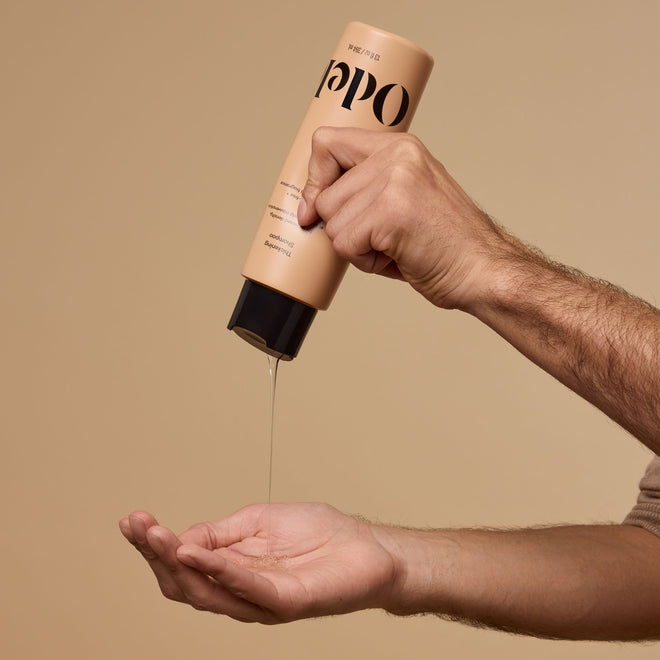 A male hand dispenses Odele Thickening Shampoo onto an outstretched palm against a plain beige background.