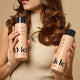 A woman with wavy brown hair holds Odele Thickening Shampoo and Conditioner against a beige background.
