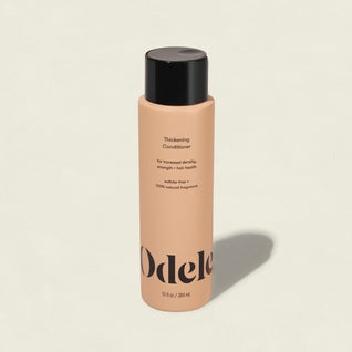 A peach-colored bottle of Odele Thickening Conditioner with a black cap stands upright on a plain background. The label promotes hair strength, density, and addresses thinning hair.
