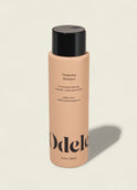 A peach-colored bottle of Odele Thickening Shampoo with a black cap rests on a light background, its label highlighting increased density, strength, and scalp rejuvenation.