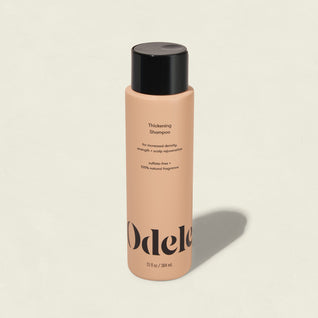 A peach-colored bottle of Odele Thickening Shampoo with a black cap rests on a light background, its label highlighting increased density, strength, and scalp rejuvenation.
