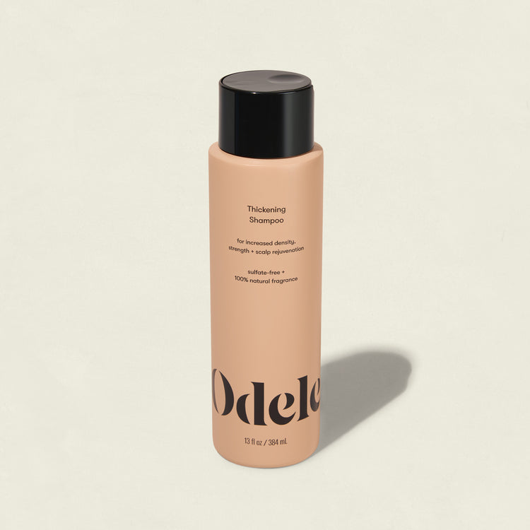 A peach-colored bottle of Odele Thickening Shampoo with a black cap rests on a light background, its label highlighting increased density, strength, and scalp rejuvenation.