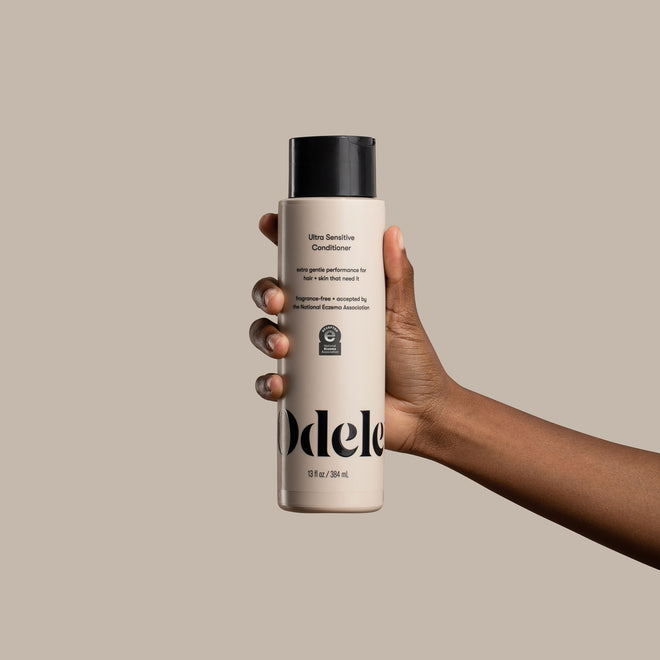 A brown skinned hand holds a bottle of Odele Ultra Sensitive Conditioner in front of a plain beige background.