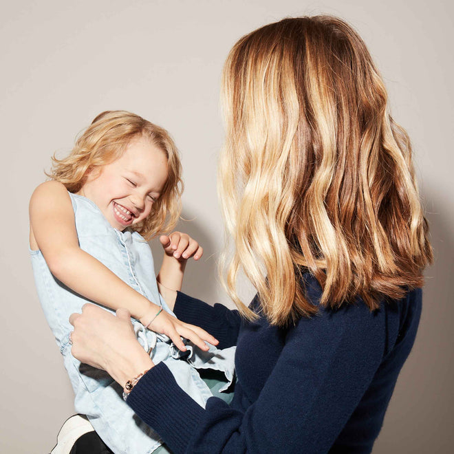 A woman with wavy blonde hair holds a laughing young girl wearing a light blue dress in front of a plain light-colored background.