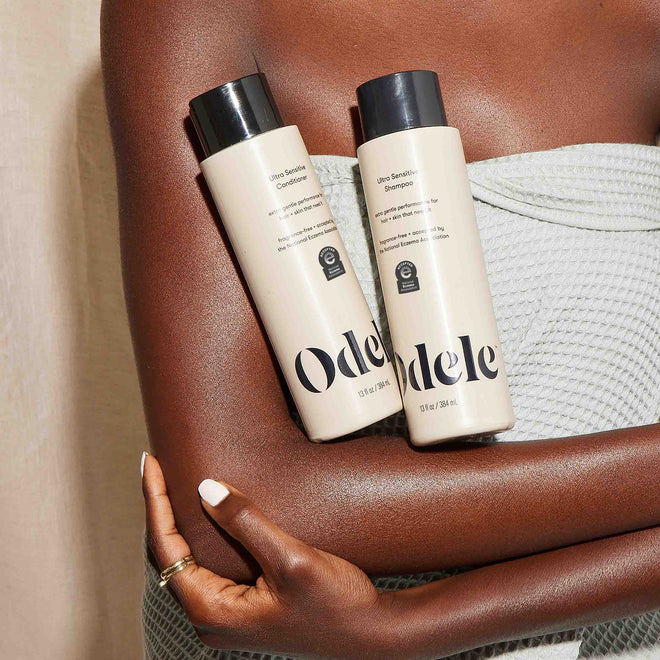 A person in a textured light gray towel holds two bottles of Odele Beauty Ultra Sensitive Shampoo, highlighting their simple beige and black design—ideal for those seeking gentle haircare for sensitive skin.
