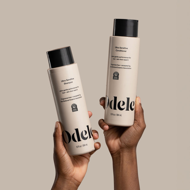 Two brown-skinned hands hold bottles of Odele Ultra Sensitive Shampoo and Conditioner in front of a plain beige background.