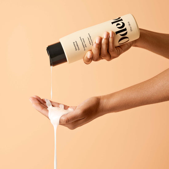 A person pours Odele Volumizing Conditioner onto their open palm against a solid peach background.