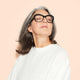 A confident older woman with gray hair and glasses, wearing a white top, poses with her head tipped upwards and eyes closed against a light peach background.